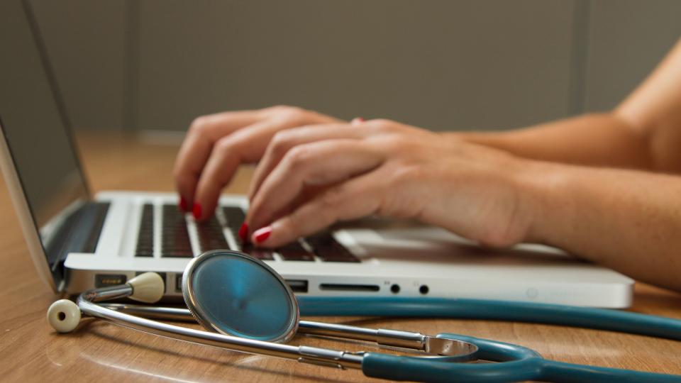 Hands type on a laptop keyboard with a stethoscope resting in photo foreground