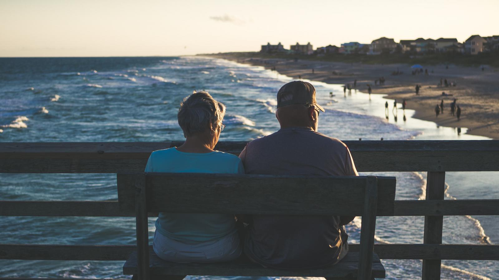 A couple sit with their backs to the camera on a bench overlooking a beach