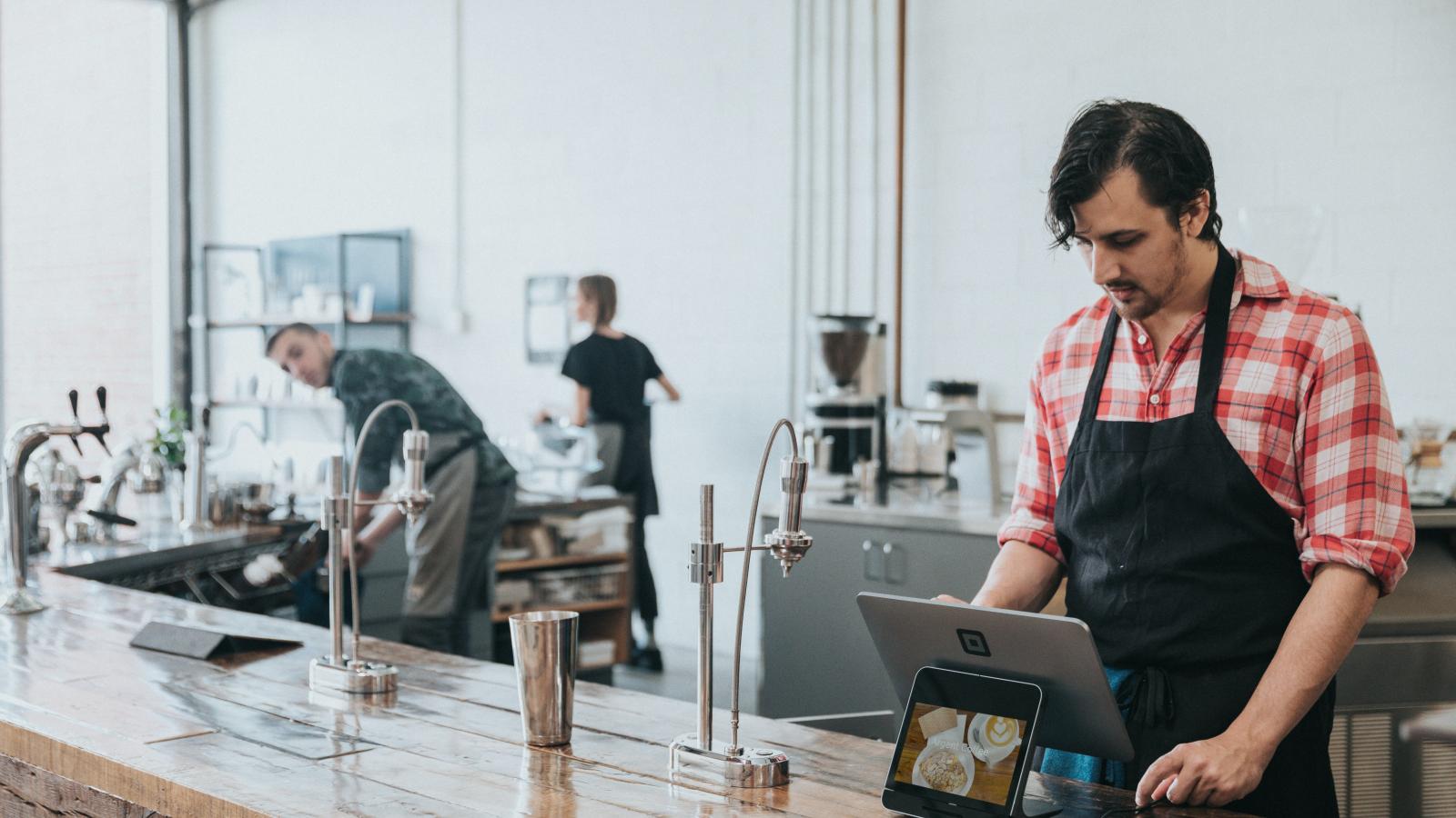 A minimum wage earner stands in an apron stands at a till in a cafe