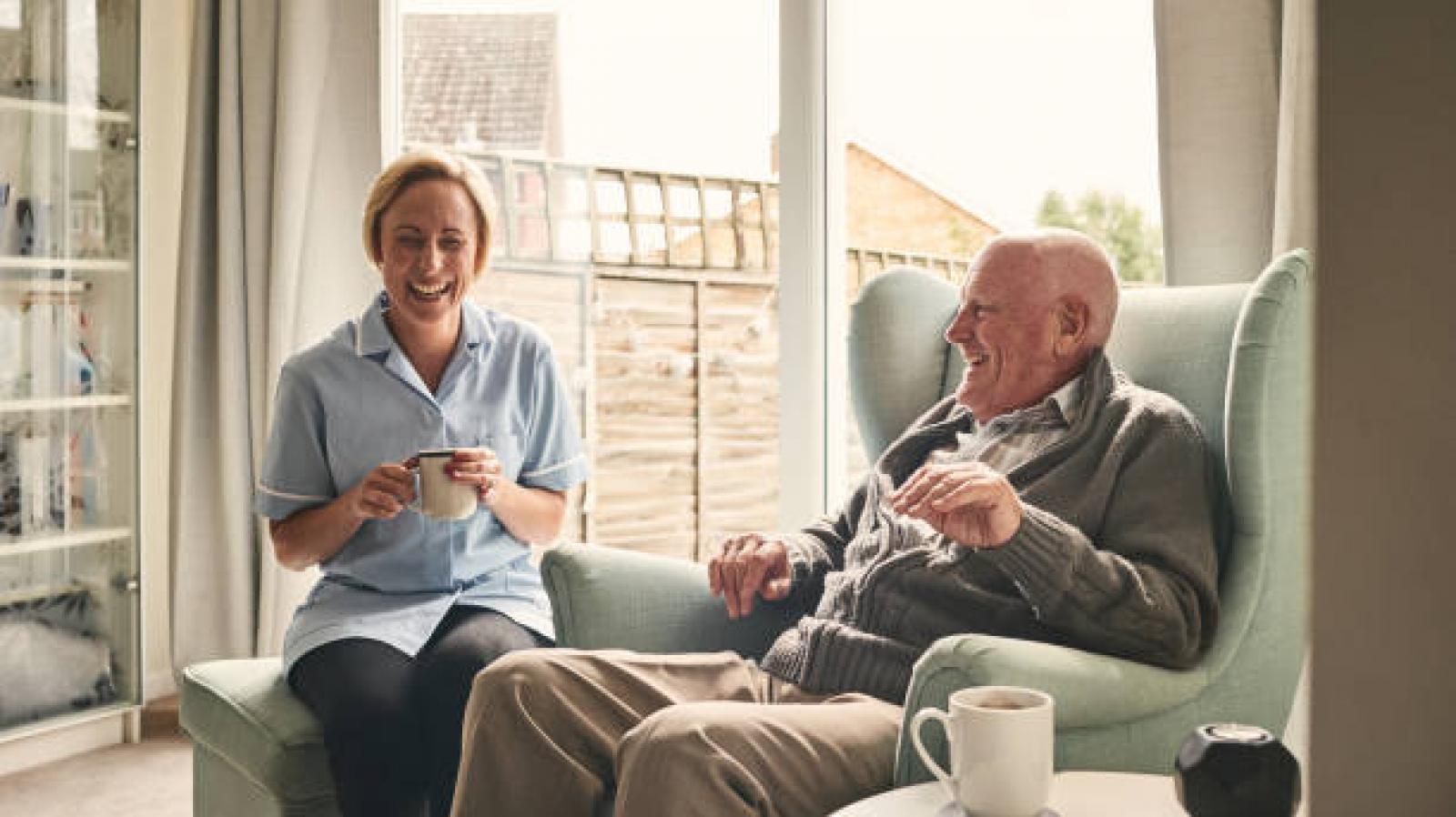 A home care nurse sits beside an elderly man whilst they drink tea