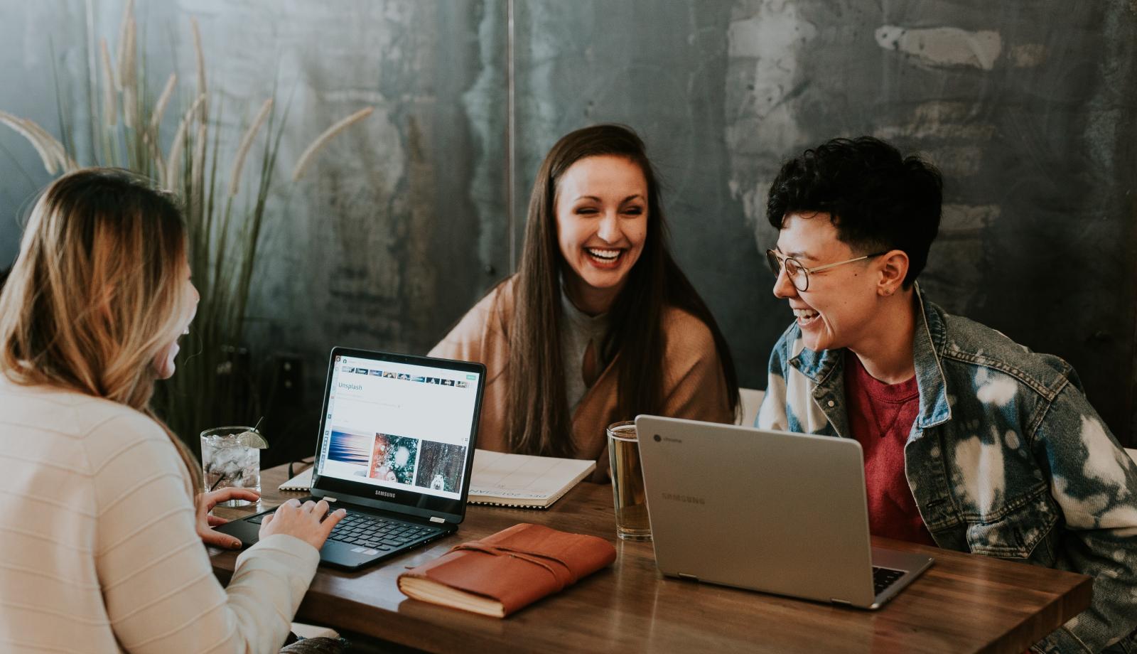 three young adults chatting at a table with their laptops open