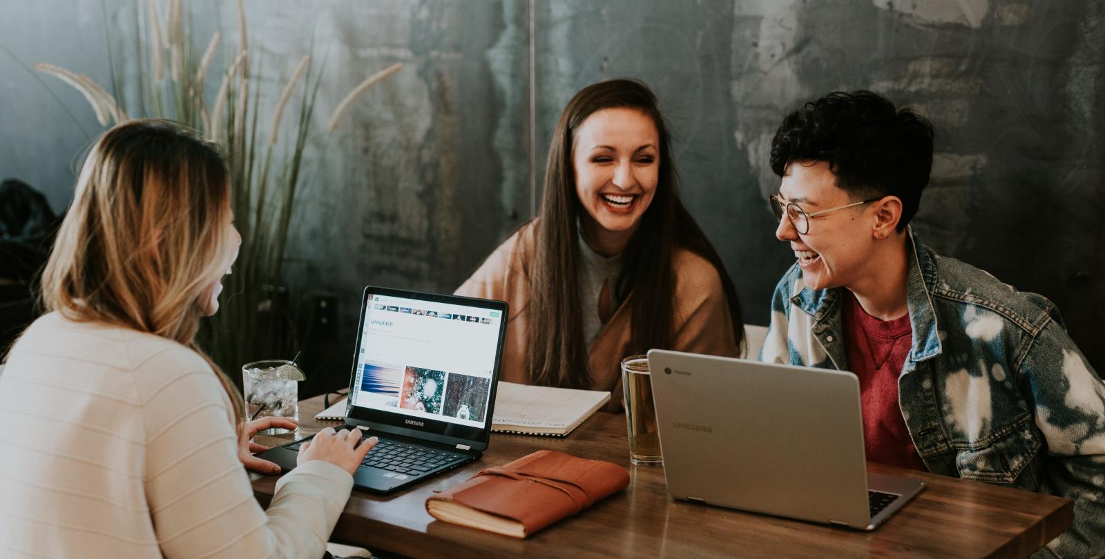 three young adults chatting and laughing at a table with their laptops open