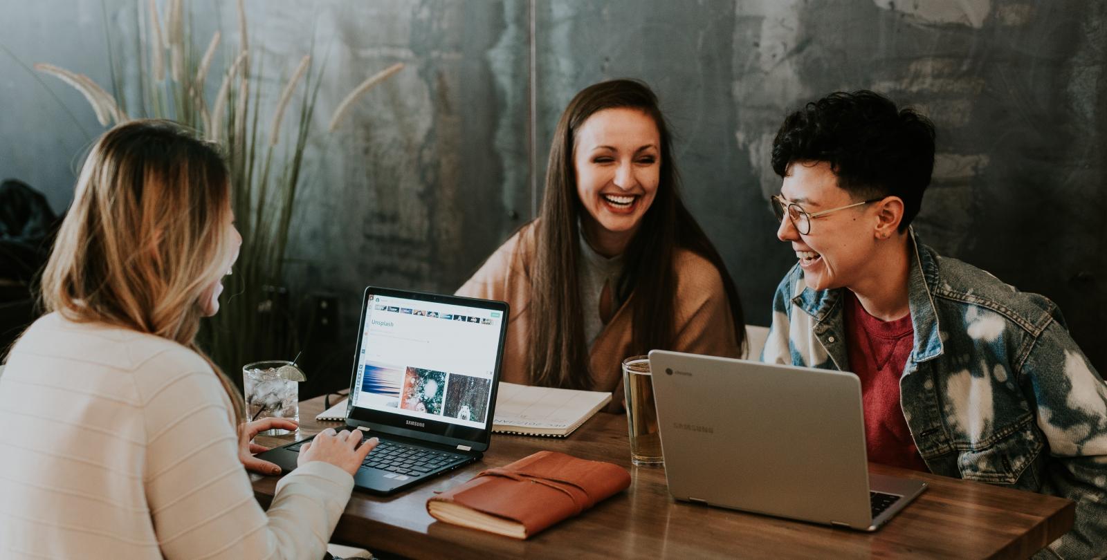 three young adults chatting and laughing at a table with their laptops open 