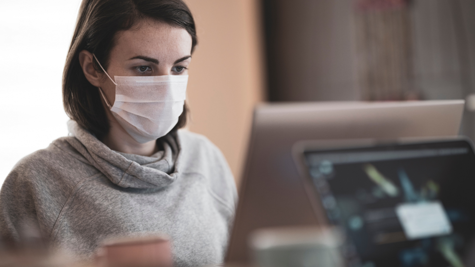 A woman in her 20s sitting at a desk wearing a face mask and looking at the computer screen