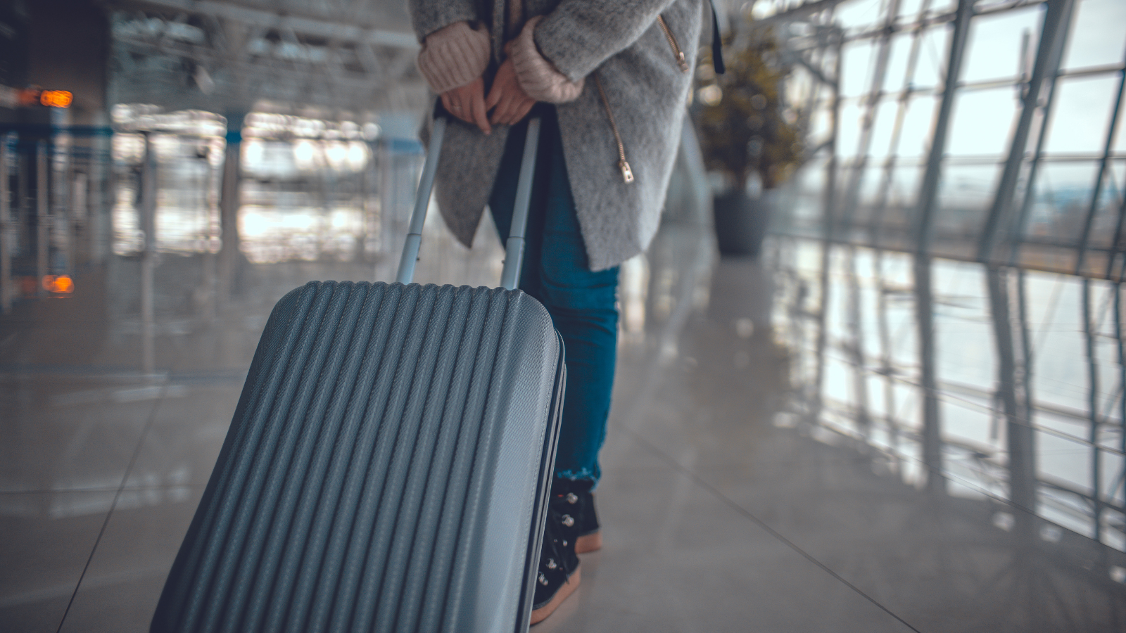 A person standing in an airport terminal, holding the handle of a gray, ribbed suitcase. The person is wearing a gray coat, blue jeans, and black shoes. The background shows large windows and reflections on the shiny floor.