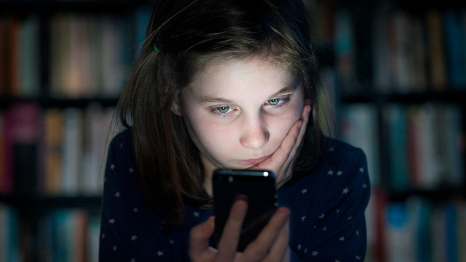A girl, around 10 years old,  holding a smartphone in front of a blurred bookshelf. She looks sad.