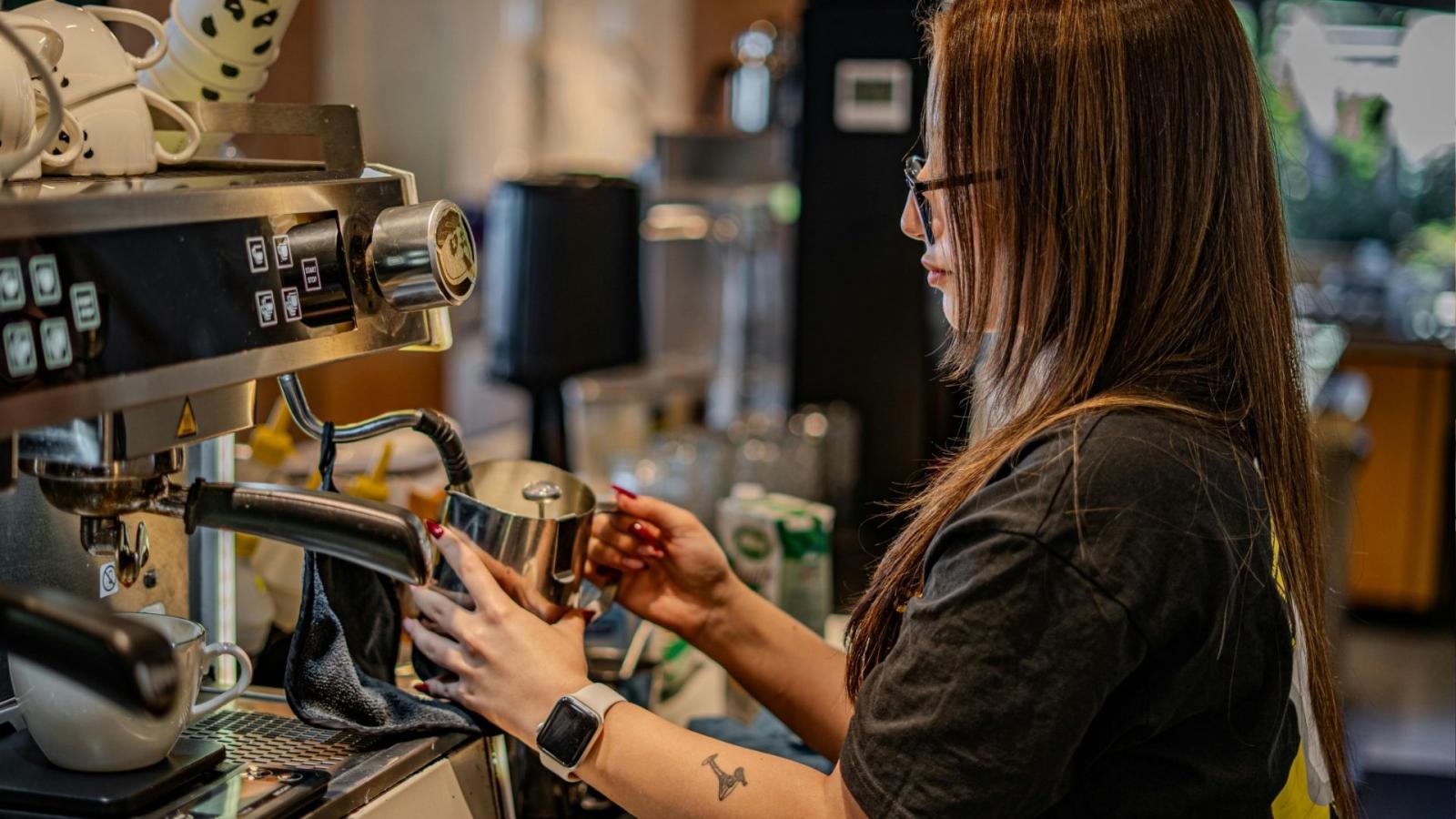 Woman with long hair and a tattoo standing at coffee machine, steaming milk