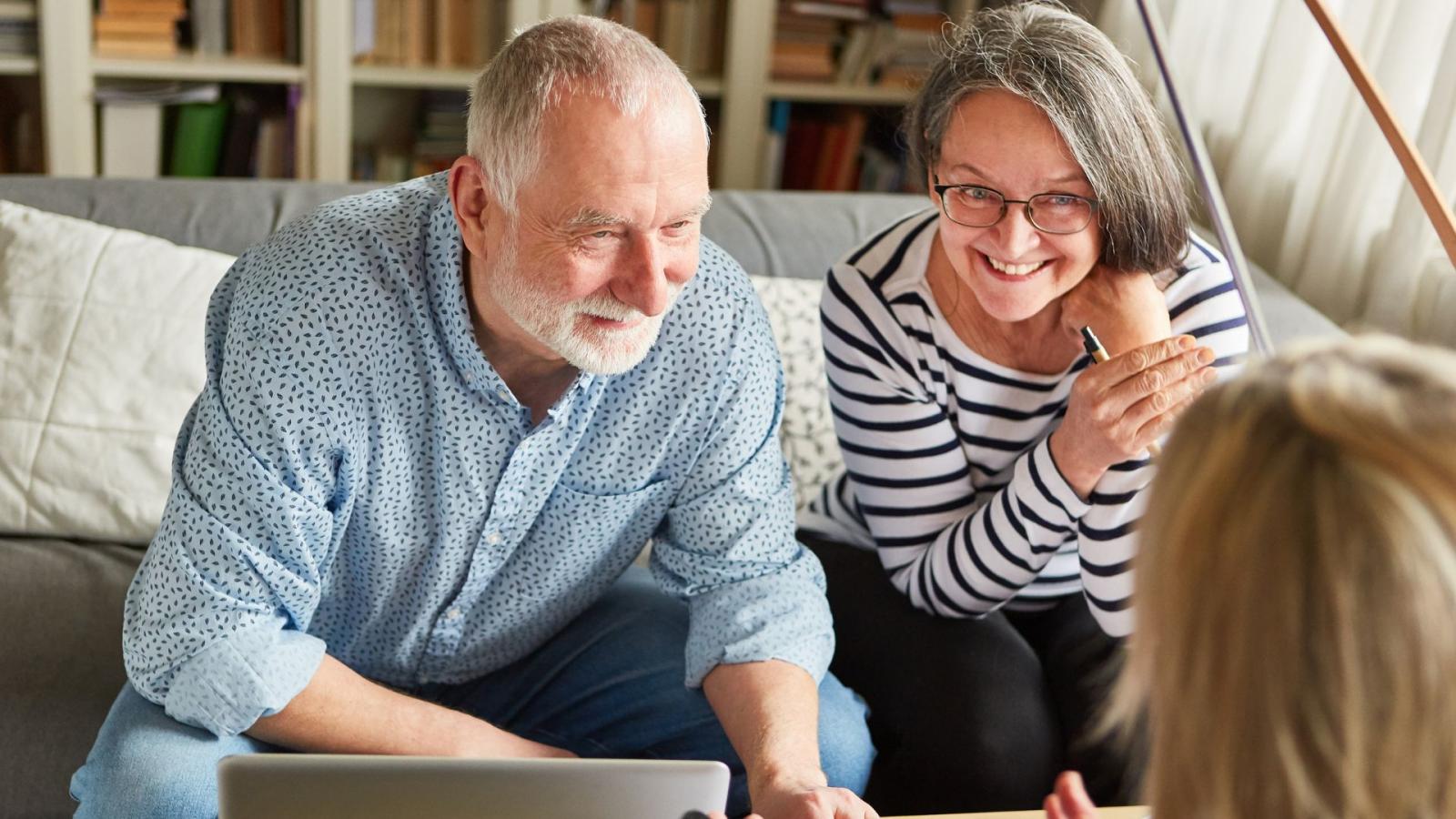 An elderly couple sits on a sofa side by side, smiling and speaking with a financial planner
