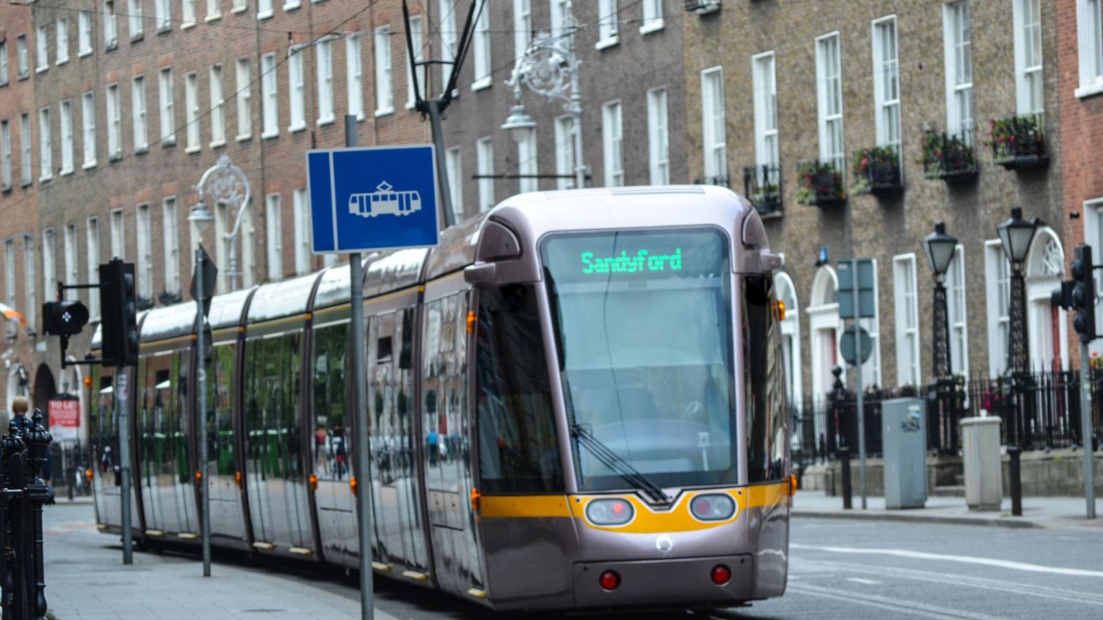 A photo of the luas in dublin city centre, with a destination of Sandyford beside a blue luas tracks sign
