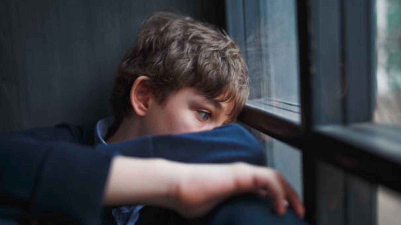 A young boy looks out a window looking sad with short brown hair