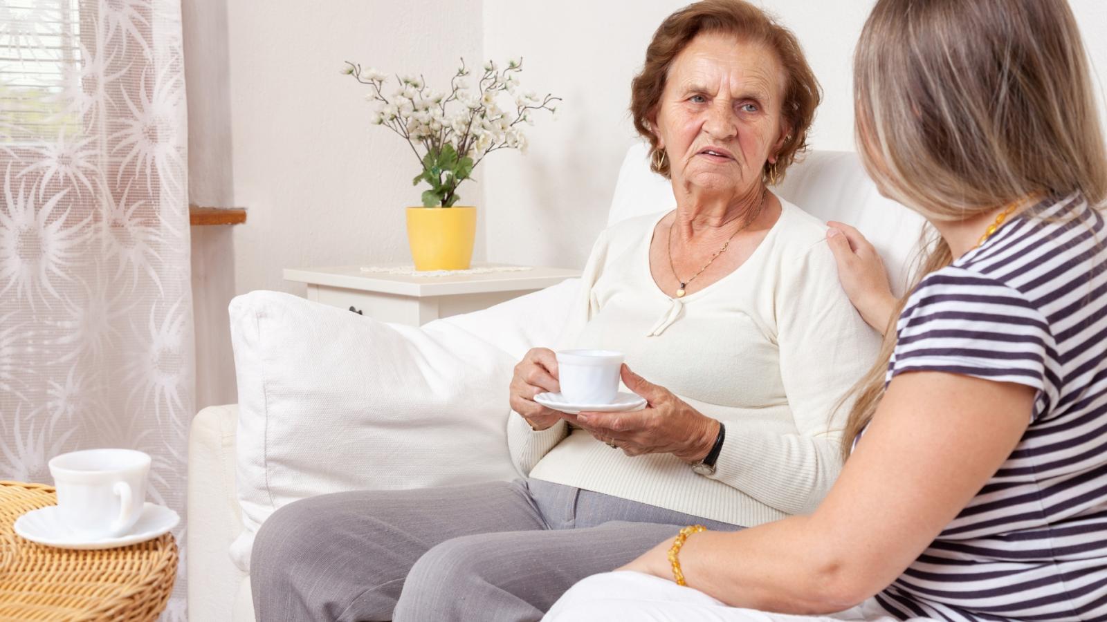 An elderly woman sits on the sofa while her family carer sits by her side, resting her hand on her shoulder